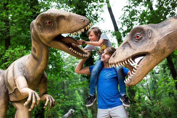 Ein Mann hat sein Kind auf den Schultern. Der Junge greift einem der beiden Dinosaurier, die neben ihnen stehen, ins Maul.