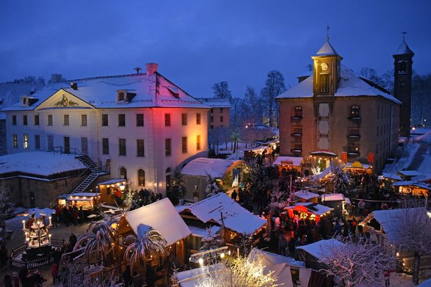 At the historic-romantic Christmas market at Königstein Fortress, there are various show booths with roofs covered in light snow. A pyramid can be seen on the left. The square with the Christmas market is surrounded by several buildings. People stand in front of the booths and look at the goods that are sold and offered.