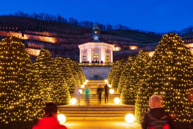 A pavilion decorated for Christmas is the center of the picture. The path uphill to the pavilion is lined with steps and decorated trees on the right and left. Five people walk up the stairs towards the pavilion.