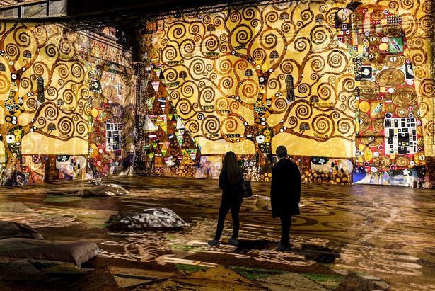 Zwei Personen schauen sich eine Ausstellung voller goldener Bilder in einem dunklen Raum an.